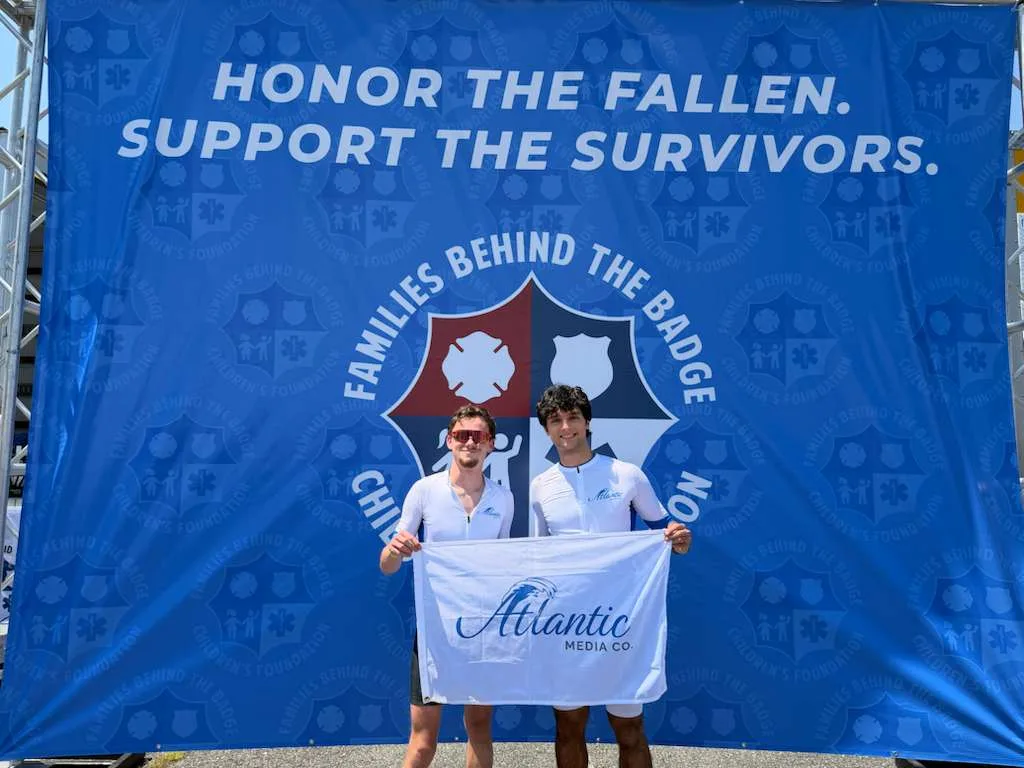 Two members of the Atlantic Media Company team holding the company flag at a Families Behind the Badge charity event, in front of an Honor the Fallen, Support the Survivors banner
