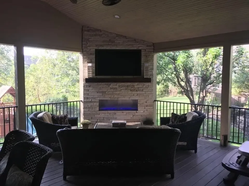 A covered deck at dusk with a linear fireplace, mounted television, and wicker seating