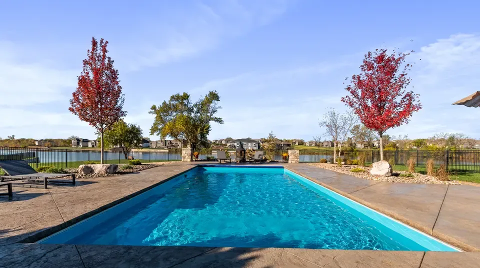A rectangular inground pool on a lakefront property, clear blue water inside clean coping with fall color in the trees behind it.