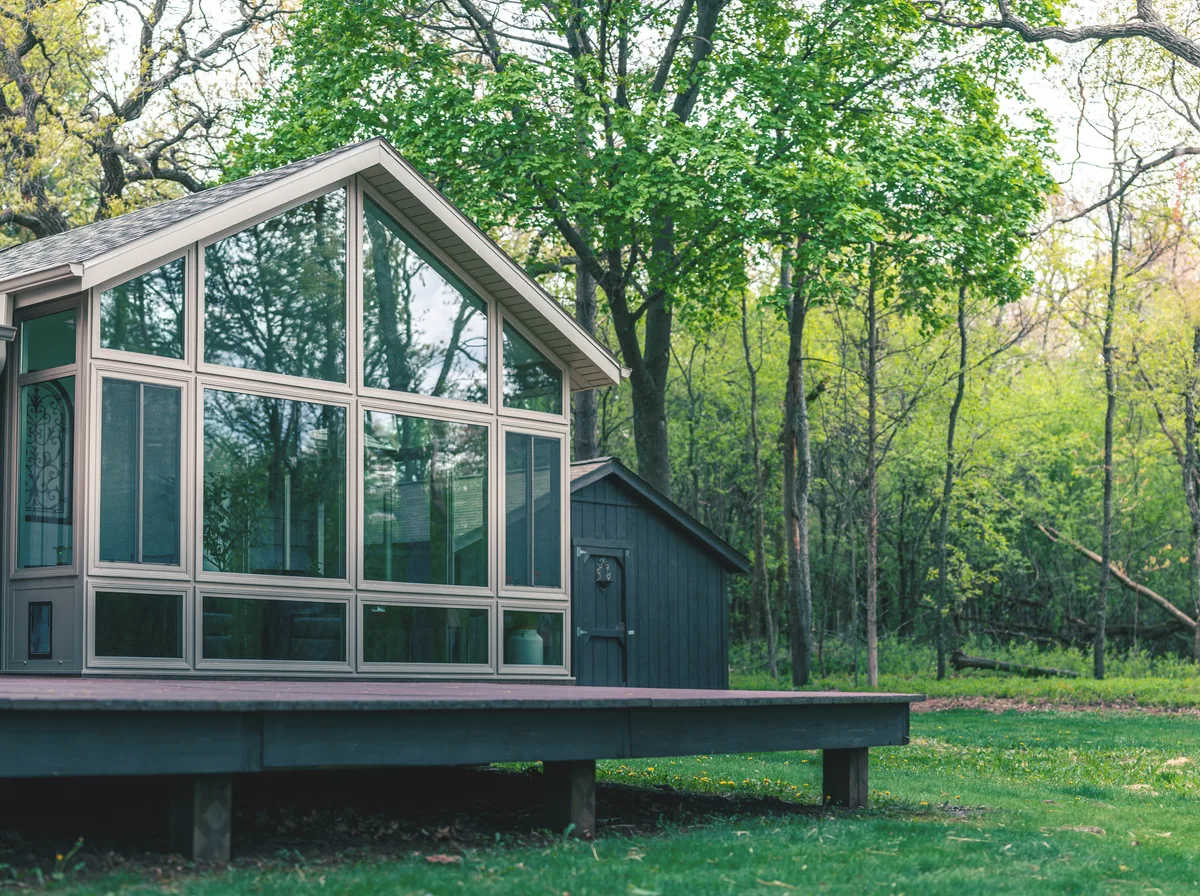 A modular, factory-built sunroom system installed on a real home, clean glass panel construction shown in understated overcast daylight with no manufacturer branding visible.