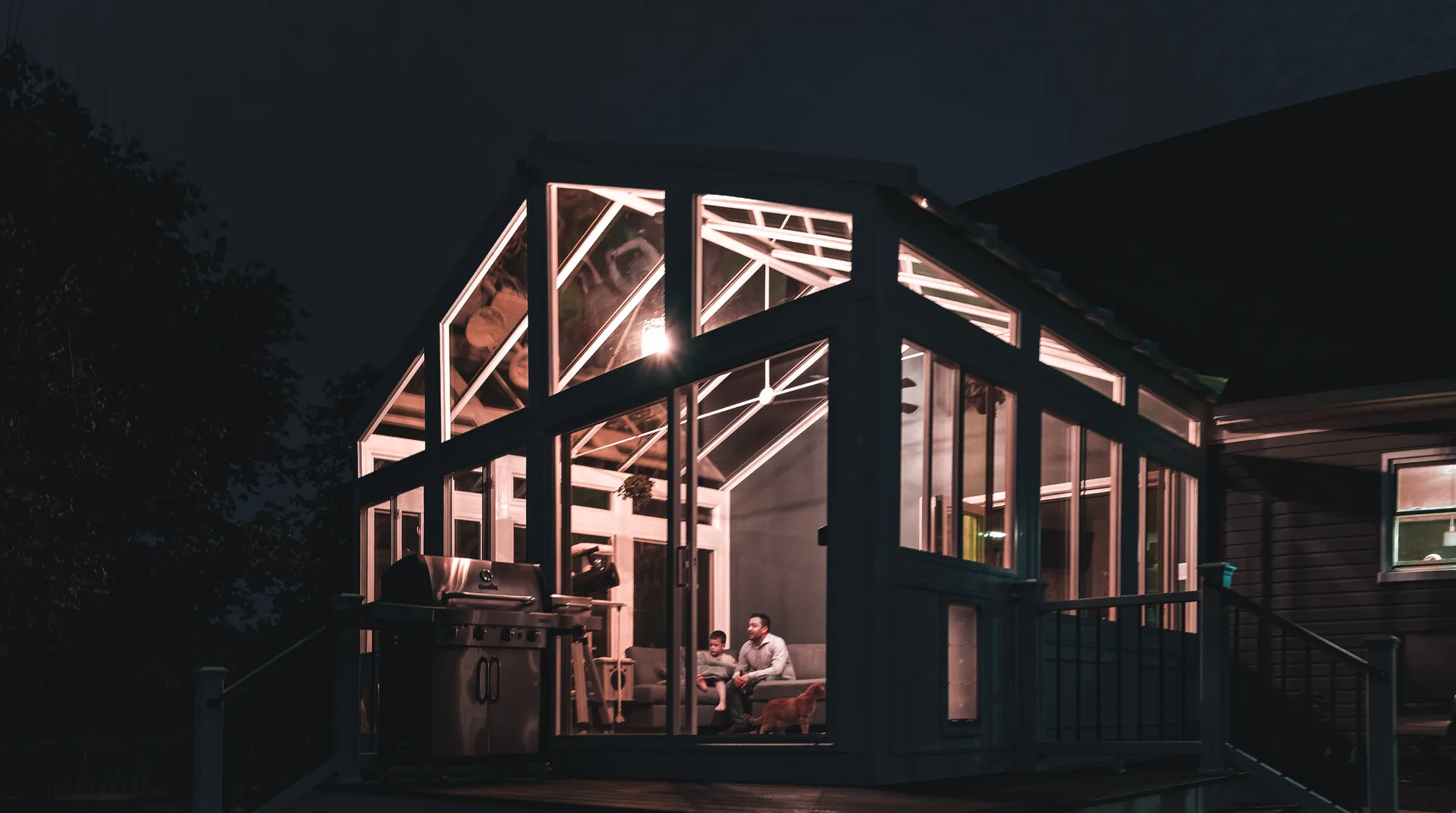 A four-season sunroom addition glowing with warm interior light at dusk, glass walls lit from within while the yard goes dark.